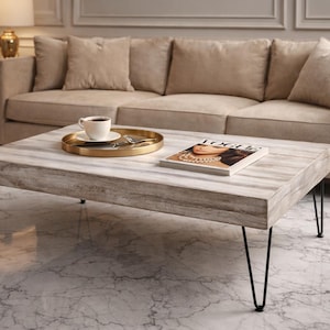 Op de afbeelding: Een rechthoekige salontafel met een lichte houtnerf afwerking en zwarte haarspeldpoten. Een gouden dienblad met een kopje koffie en een tijdschrift staan op de tafel. Een beige bank en marmeren vloer completeren de scène. De tafel is ongeveer 120 cm lang.