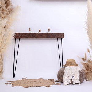 May include: A rustic wooden console table with black hairpin legs, topped with three small decorative items. The scene includes a jute rug, a hat, and a baby outfit on a round ottoman, and tall pampas grass.
