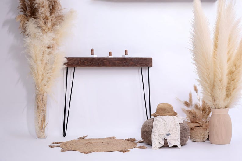 May include: A rustic wooden console table with black hairpin legs, topped with three small decorative items. The scene includes a jute rug, a hat, and a baby outfit on a round ottoman, and tall pampas grass.