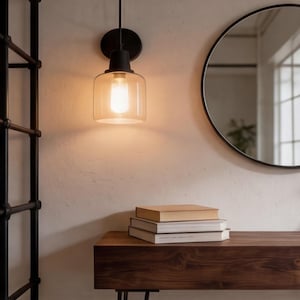 Puede incluir: Una l&aacute;mpara de pared negra con una pantalla de vidrio transparente emite un brillo c&aacute;lido y acogedor. Debajo, una mesa de madera oscura sostiene una pila de libros. Un espejo redondo con marco negro cuelga de la pared, reflejando la luz natural.