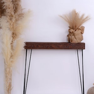 Può includere: Una consolle in legno marrone scuro con gambe a forcina nere. Il tavolo è decorato con un fascio di erba della pampa essiccata in un sacco di juta e un tappeto rotondo in juta. Lo sfondo è bianco.