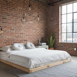 Pode incluir: Um quarto moderno com uma cama baixa de madeira, roupa de cama branca e almofadas decorativas redondas. O quarto apresenta uma parede de tijolo, uma janela grande e luminárias de estilo industrial. Um tapete cinza e plantas em vasos completam a decoração.