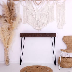May include: A room interior featuring a dark wood console table with black hairpin legs, a woven chair, and a woven basket. A large vase of pampas grass and a macrame wall hanging add to the bohemian decor.