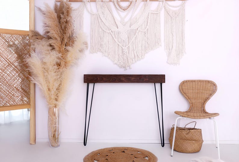 May include: A room interior featuring a dark wood console table with black hairpin legs, a woven chair, and a woven basket. A large vase of pampas grass and a macrame wall hanging add to the bohemian decor.