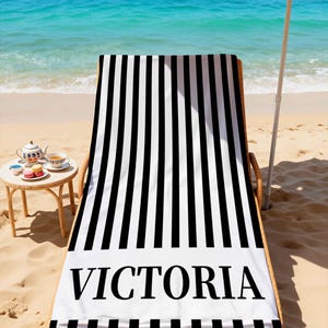 May include: A black and white striped beach towel with the name "VICTORIA" printed in large, bold letters. The towel is draped over a wicker beach chair on a sandy beach, with the ocean and a beach umbrella in the background.