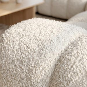 Op de afbeelding: Een cr&egrave;mekleurige bank met een dikke, getextureerde boucl&eacute; stoffering. Het modulaire ontwerp bestaat uit afgeronde, pluche secties die een zacht, onregelmatig oppervlak cre&euml;ren over het gehele zitgedeelte van het meubelstuk.