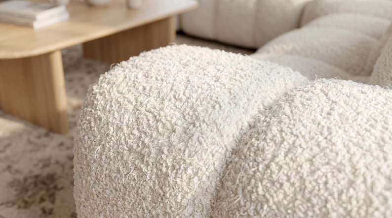 Op de afbeelding: Een cr&egrave;mekleurige bank met een dikke, getextureerde boucl&eacute; stoffering. Het modulaire ontwerp bestaat uit afgeronde, pluche secties die een zacht, onregelmatig oppervlak cre&euml;ren over het gehele zitgedeelte van het meubelstuk.
