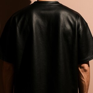 May include: A black, short-sleeved shirt made of a leather-like material. The shirt has a crew neck and a loose fit. The image is a close-up of the back of the shirt, with the shoulders and upper arms visible. The background is a solid, neutral color.