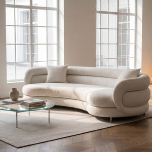 May include: A curved, off-white sofa with rounded, layered armrests and two matching throw pillows. In front of the sofa sits a round, clear glass coffee table with thin metal legs, holding a small ceramic vase and two stacked books.