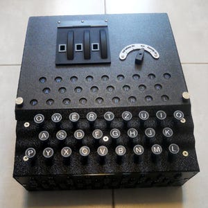Könnte beinhalten: Eine schwarze Enigma-Maschinen-Nachbildung mit QWERTZ-Tastaturlayout, runden Tasten, einem Lampenfeld mit Buchstaben, drei Rotoren an der Oberseite und einem Schalter mit deutschem Text. Das Gerät verfügt über eine strukturierte Oberfläche.