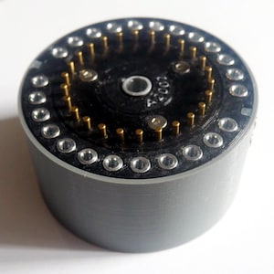 Puede incluir: Un componente electrónico cilíndrico con un exterior gris liso y una superficie superior negra. La parte superior presenta una disposición circular de orificios con borde plateado, un anillo interior de pines dorados y un puerto central plateado con la etiqueta A2007.