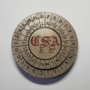 Può includere: Una ruota di cifratura rotonda, nei toni del beige e del marrone, con lettere incise sul bordo e un disco centrale con la scritta "USA" in rosso. La ruota è progettata per codificare e decodificare messaggi.
