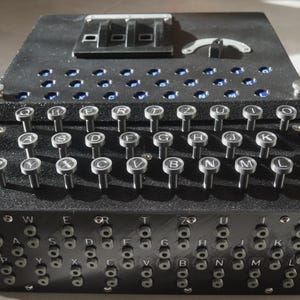 Puede incluir: Una máquina Enigma negra con un teclado de teclas con letras y una serie de interruptores y diales. La máquina tiene forma rectangular con una superficie texturizada, probablemente una réplica.