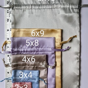 May include: A set of six satin drawstring bags in various sizes, ranging from 2x3 inches to 8x13 inches. The bags are arranged in order of size, with the largest bag at the top and the smallest bag at the bottom. The bags are in the following colors: light gray, gold, lavender, brown, light blue, and pink. The text "Sizes are not available in every color." is printed at the top of the image. The text "8x13", "6x9", "5x8", "4x6", "3x4", and "2x3" is printed on each bag, indicating the size of the bag in inches.