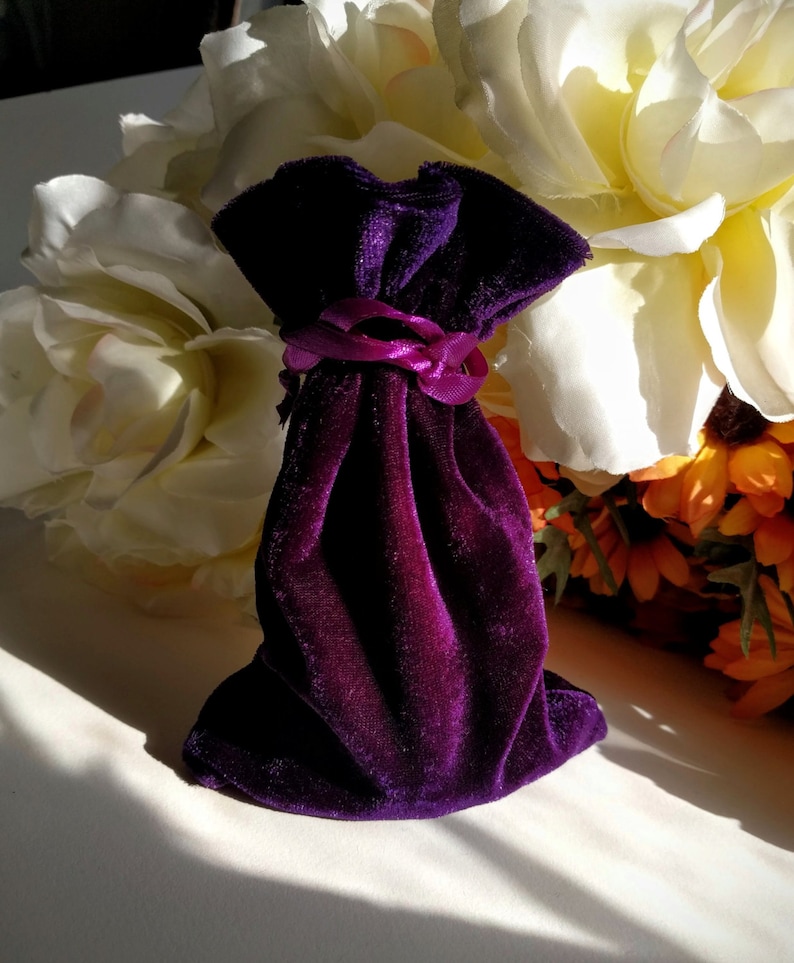 May include: A small purple velvet pouch with a pink ribbon tied around the top. The pouch is sitting on a white surface with white flowers in the background.