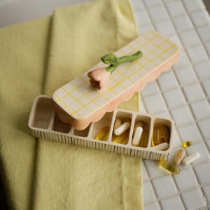 May include: A ceramic pill organizer with a pink and yellow checkered lid, topped with a pink flower and green stem. The open compartment reveals pills. The word "SUN" is visible on the organizer.