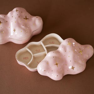 Pode incluir: Dois porta-comprimidos de cerâmica em forma de nuvem, em rosa claro com detalhes de estrelas e pontos dourados. Um recipiente está fechado, enquanto o outro está aberto, revelando quatro compartimentos internos com o texto MON, TUE, WED e THU.