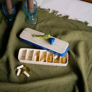 May include: A ceramic pill organiser with a blue and white design, featuring a blue flower accent on the lid. The organiser is open, revealing compartments labelled for each day of the week, filled with pills. The background is a green fabric.