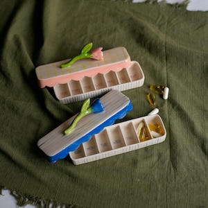 May include: Two ceramic pill organisers with floral accents. One has a pink scalloped edge and a pink flower, the other has a blue scalloped edge and a blue flower. Each has compartments labelled for the days of the week.