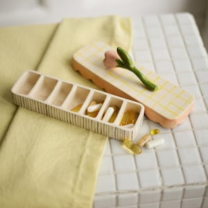 May include: A beige ceramic pill organiser with seven compartments, filled with pills. A matching lid with a pink flower and green stem rests nearby. The organiser is on a white tile surface with a yellow cloth.