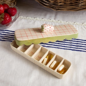 May include: A rectangular ceramic pill organiser with seven compartments labelled with the days of the week. The lid features a red grid pattern on a cream background with a sculpted strawberry handle and a scalloped light green edge.