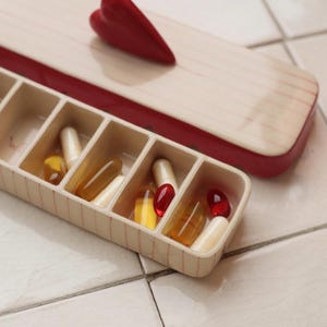 May include: A cream-colored pill organizer with a red trim and a red heart-shaped accent. The organizer is filled with various capsules and pills in shades of yellow, red, and white. The lid has a striped pattern.