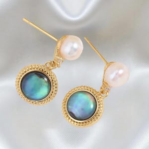 May include: A pair of gold-colored earrings featuring a pearl stud and a dangling circular pendant. The pendant has a gold-colored frame with a textured edge, surrounding a shimmering, iridescent blue-green stone.