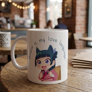 May include: White ceramic mug featuring a cartoon anime character with teal hair and a pink outfit. The mug has the text "Poison is my love language" in a whimsical font. The mug is on a wooden table.