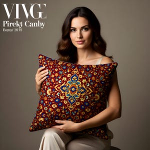 May include: A decorative pillow with a burgundy background and a detailed floral pattern in shades of blue, yellow, and orange. The pillow has a white border and is held by a person. The text "VIVE" and "Pirekt Canby" are visible.