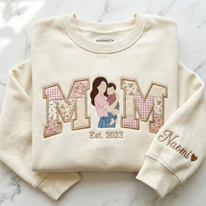 May include: Cream-colored sweatshirt with the word "MOM" in a patchwork design, featuring floral and gingham patterns. An illustration of a mother and child is in the center. The sleeve has the name "Naomi" embroidered with a heart.