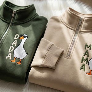 May include: Two quarter-zip sweatshirts, one olive green and one beige. Each features an embroidered goose wearing sunglasses. The green sweatshirt has the word "DADA" embroidered next to the goose, while the beige one says "MAMA". Both have ribbed cuffs and collars.