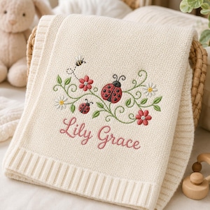 May include: A cream-coloured knit blanket featuring an embroidered design of a red ladybird, a smaller ladybird, a bee, green vines, and flowers. The name Lily Grace is embroidered in pink cursive script below the floral and insect pattern.