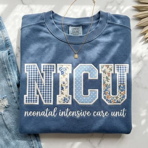 May include: Blue sweatshirt with "NICU" in white, floral, and gingham patterns. The words "neonatal intensive care unit" are below the letters. A gold necklace with a small pendant is on the sweatshirt. Blue jeans are on the left.