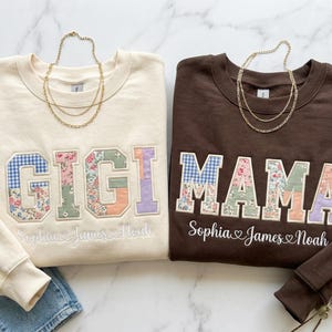 May include: Two sweatshirts, cream and brown, with floral and patterned lettering. The cream sweatshirt says "GIGI" and the brown one says "MAMA". Both have embroidered names below. Gold necklaces are draped over the sweatshirts.
