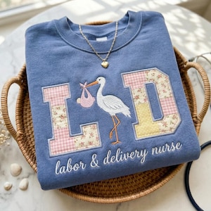 May include: A blue sweatshirt with the embroidered letters "L D" and a stork carrying a baby. The letters are decorated with floral patterns. The text "labor & delivery nurse" is embroidered below the letters.