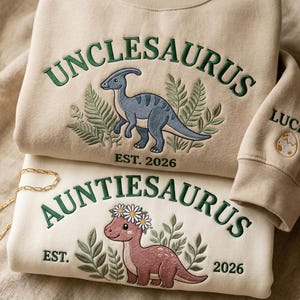 May include: Two beige and cream sweatshirts with dinosaur embroidery. One says "UNCLESAURUS" with a blue dinosaur and "EST. 2026". The other says "AUNTIESAURUS" with a pink dinosaur and "EST. 2026". A gold chain is visible.