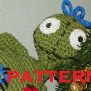 May include: A green crocheted stuffed creature with large white eyes featuring black button pupils, a blue yarn tuft on its head, and blue and yellow circular patches on its body. The word PATTERN is printed in red across the centre of the image.