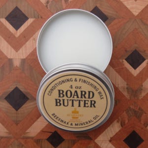 May include: A 4 oz. tin of Board Butter wood conditioning and finishing wax, with the lid slightly ajar. The label reads "Conditioning & Finishing Wax" and "Beeswax & Mineral Oil". The background is a wooden surface with a geometric pattern.