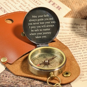 Puede incluir: Una brújula de estilo vintage con acabado de latón, abierta para revelar la esfera de la brújula y un mensaje. La brújula está alojada en un estuche de cuero marrón. El texto en la tapa dice: "Que tu fe te guíe siempre..."