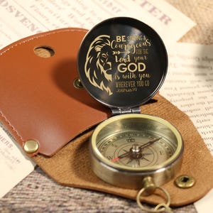 May include: A vintage-style compass with a leather case. The compass has a black lid with a lion graphic and the text "BE STRONG & COURAGEOUS for the Lord your GOD is with you WHEREVER YOU GO Joshua 1:9".
