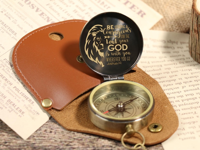 May include: A vintage-style compass with a leather case. The compass has a black lid with a lion graphic and the text "BE STRONG & COURAGEOUS for the Lord your GOD is with you WHEREVER YOU GO Joshua 1:9".