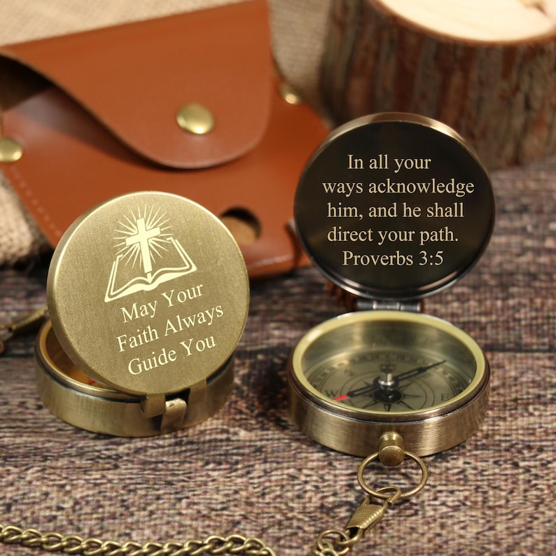 May include: Two antique brass compasses with inspirational quotes engraved on the lids. One reads "May Your Faith Always Guide You" and the other "In all your ways acknowledge him, and he shall direct your path. Proverbs 3:5". A brown leather case is in the background.