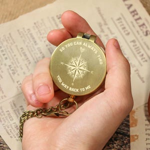 May include: A round, brass compass with a chain is held in a hand. The compass face features a compass rose design and the engraved text: "SO YOU CAN ALWAYS FIND YOU WAY BACK TO ME." The background includes vintage paper and a leather case.