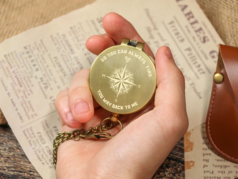 May include: A round, brass compass with a chain is held in a hand. The compass face features a compass rose design and the engraved text: "SO YOU CAN ALWAYS FIND YOU WAY BACK TO ME." The background includes vintage paper and a leather case.