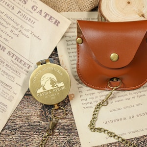 May include: A personalized brass compass with a mountain and sun design, engraved with "FOR OUR ADVENTURES" and a name and date. It includes a brown leather pouch and a chain. The compass is on a vintage paper background.
