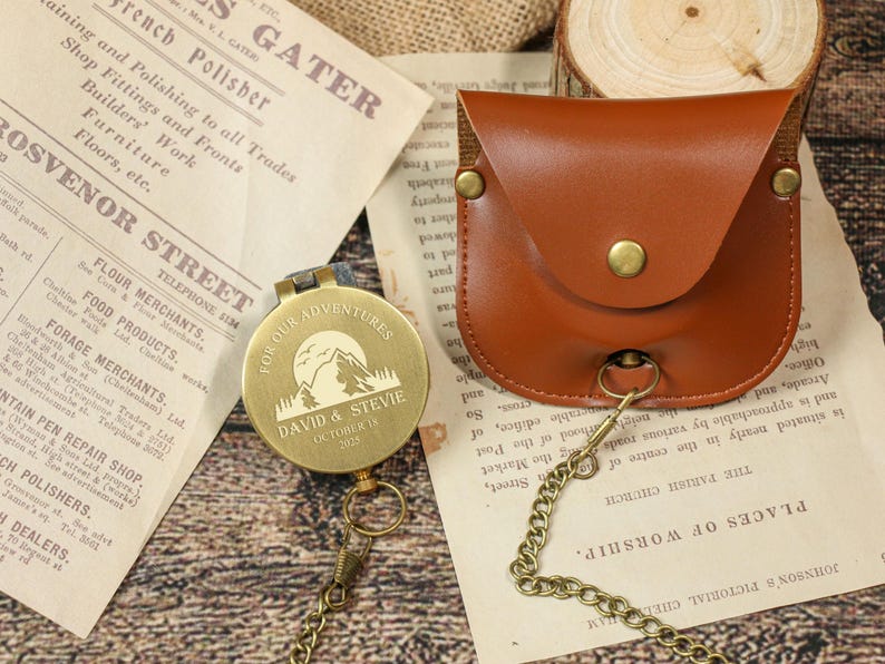 May include: A personalized brass compass with a mountain and sun design, engraved with "FOR OUR ADVENTURES" and a name and date. It includes a brown leather pouch and a chain. The compass is on a vintage paper background.
