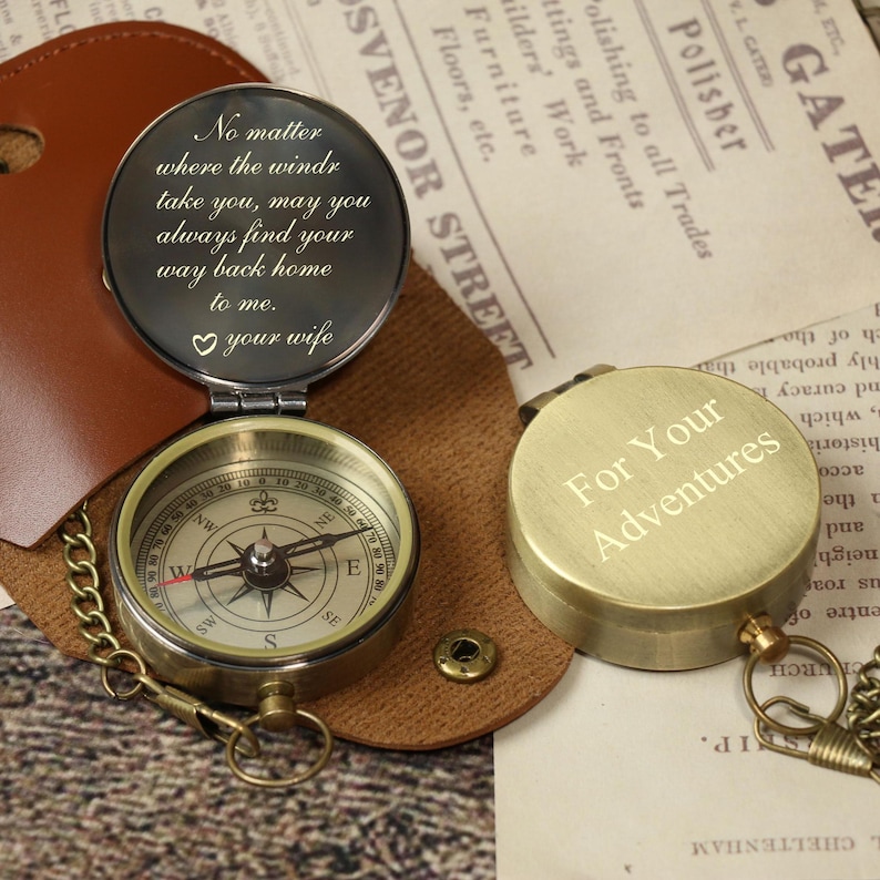 May include: A vintage-style compass with a brass finish, open to reveal the compass face and a lid with an engraved message. The compass is in a brown leather case. The closed compass has the text "For Your Adventures".