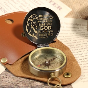 May include: A vintage-style compass with a brass finish, open to reveal the compass dial and a black lid with a lion graphic and the text "BE STRONG & COURAGEOUS for the Lord your GOD is with you WHEREVER YOU GO Joshua 1:9". The compass is in a brown leather case.