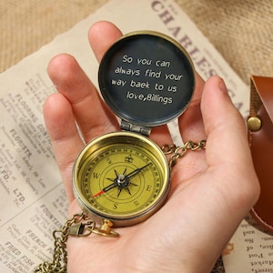 May include: A vintage brass compass with a gold-colored face and a chain, held open to reveal an inscription. The compass is set against a backdrop of aged paper and a leather pouch.