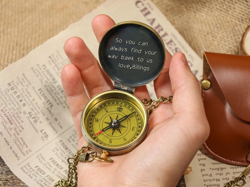 May include: A vintage brass compass with a gold-colored face and a chain, held open to reveal an inscription. The compass is set against a backdrop of aged paper and a leather pouch.